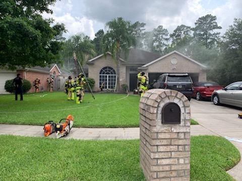 Firefighters in yellow suits gather outside a smoking brick house with a hose, preparing equipment. Two cars are parked in the driveway. A mailbox is in the foreground. Lush green lawn and surrounding trees are visible under a cloudy sky.