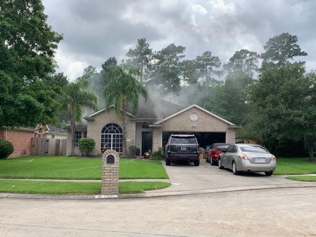 A suburban house with gray smoke rising from the roof. Several cars are parked in the driveway. The grass is green and trees surround the house under a cloudy sky.