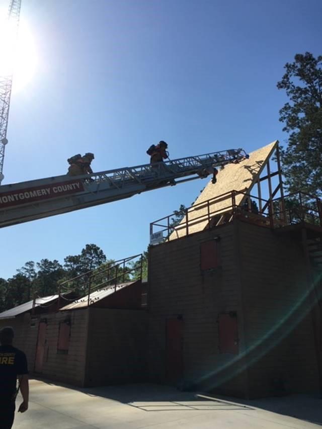 Firefighters climb an extended fire truck ladder to reach the sloped roof of a building. The scene is outdoors during daytime, with trees and a clear sky in the background.