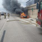 Firefighters respond to a car engulfed in flames on a highway. Thick black smoke rises, while a fire truck is parked nearby. Firefighters approach the scene, unrolling hoses to extinguish the fire. Traffic is visible on the opposite side of the road.