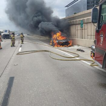 Firefighters respond to a car engulfed in flames on a highway. Thick black smoke rises, while a fire truck is parked nearby. Firefighters approach the scene, unrolling hoses to extinguish the fire. Traffic is visible on the opposite side of the road.