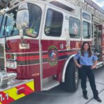 A firefighter stands beside a large red fire truck, smiling. The truck has visible decals and equipment on it. The sky is partly cloudy, and the ground is paved.
