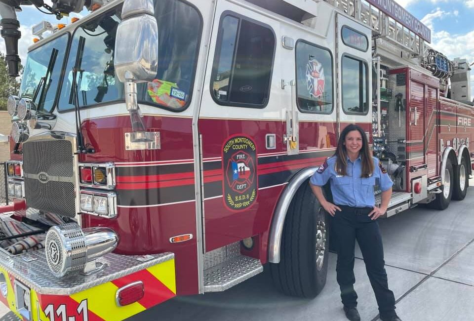 A firefighter stands beside a large red fire truck, smiling. The truck has visible decals and equipment on it. The sky is partly cloudy, and the ground is paved.