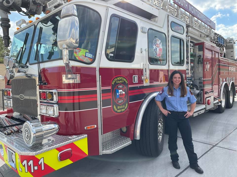A firefighter stands beside a large red fire truck, smiling. The truck has visible decals and equipment on it. The sky is partly cloudy, and the ground is paved.