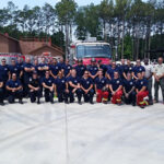 A large group of firefighters in uniform pose for a photo in front of a fire truck. Trees and buildings are visible in the background. Some are wearing navy shirts, while others have red or yellow pants. Everyone is smiling.