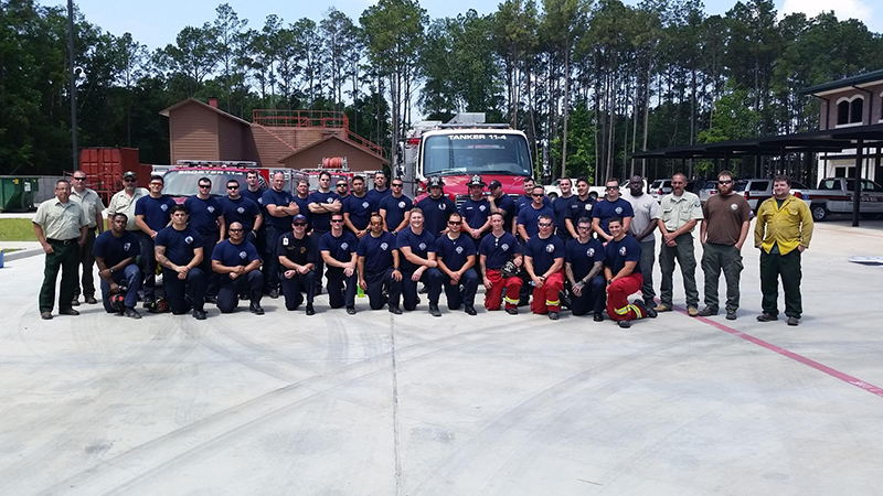 A large group of firefighters in uniform pose for a photo in front of a fire truck. Trees and buildings are visible in the background. Some are wearing navy shirts, while others have red or yellow pants. Everyone is smiling.