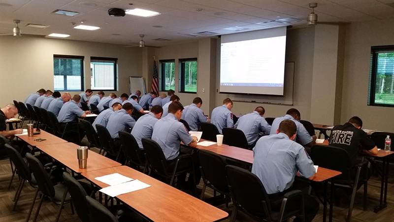 A group of people in blue uniforms sit at desks in a classroom, focused on writing. A presentation is displayed on a screen at the front of the room. An American flag is in the corner. The room has several windows and ceiling lights.