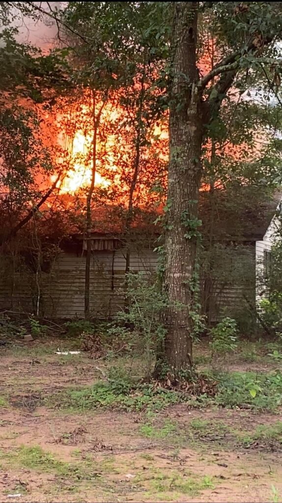 A house engulfed in flames is seen through the trees, with intense orange and yellow flames rising above the roof. The surrounding area has several trees and bushes, with a clear patch of ground in the foreground.