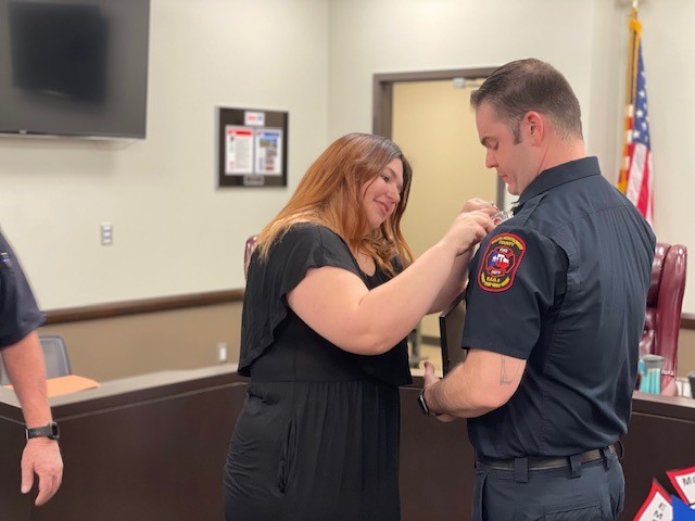 A woman in a black dress pins a badge on a firefighters uniform in an office setting. An American flag is visible in the background. They both appear focused and engaged in the moment.