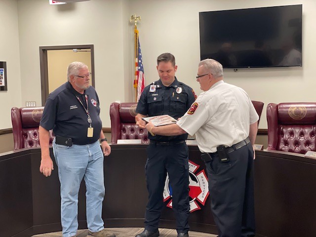 Three men, two in fire department uniforms, stand in a meeting room. The middle man receives a plaque from the man on the right. An American flag and a large TV screen are in the background.
