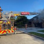 Firefighters respond to a house fire, with smoke billowing from the roof. A fire truck with bright red and yellow markings has its ladder extended, and firefighters, in full gear, are positioned on and around it in a suburban neighborhood.
