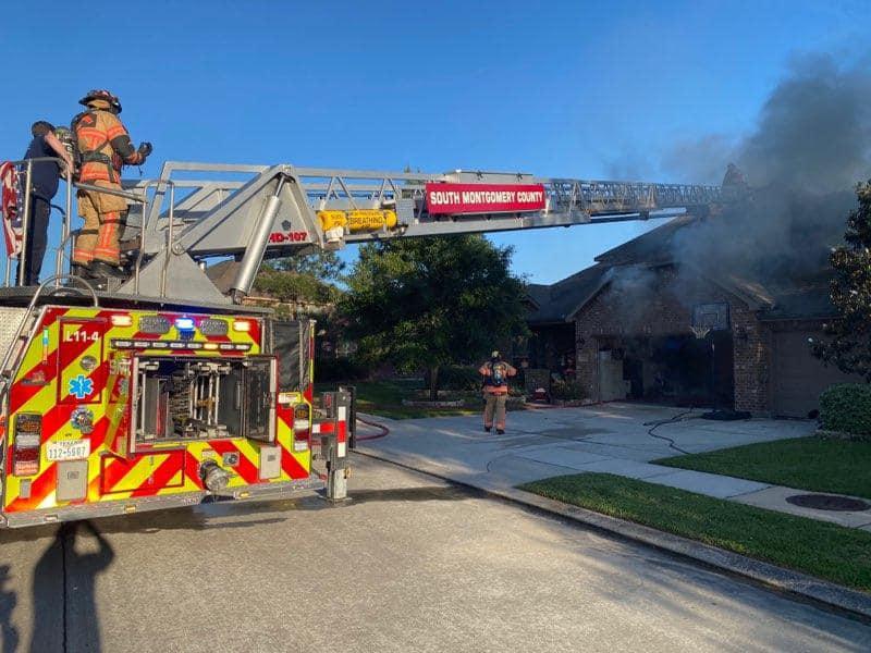 Firefighters respond to a house fire, with smoke billowing from the roof. A fire truck with bright red and yellow markings has its ladder extended, and firefighters, in full gear, are positioned on and around it in a suburban neighborhood.