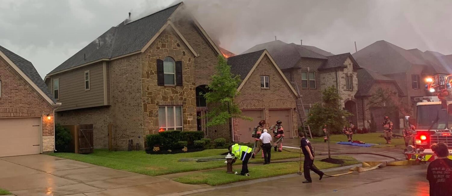A house fire scene shows smoke rising from a suburban home under a gray sky. Firefighters and emergency personnel are on site, with a fire truck parked beside the house. Several people stand on the wet street, observing the situation.