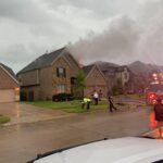 A house fire scene shows smoke rising from a suburban home under a gray sky. Firefighters and emergency personnel are on site, with a fire truck parked beside the house. Several people stand on the wet street, observing the situation.