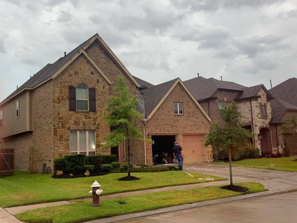 A two-story brick house with a stone facade, surrounded by a neatly manicured lawn and trees. A person stands in the driveway near the garage. A fire hydrant is visible on the grass by the sidewalk. The sky is overcast.