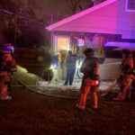 Firefighters in gear stand around the side of a house at night. Some hold hoses, and others appear to survey damage near a window. The scene is illuminated by red and purple lights. The ground is wet, suggesting firefighting efforts.