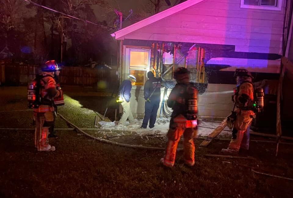 Firefighters in gear stand around the side of a house at night. Some hold hoses, and others appear to survey damage near a window. The scene is illuminated by red and purple lights. The ground is wet, suggesting firefighting efforts.