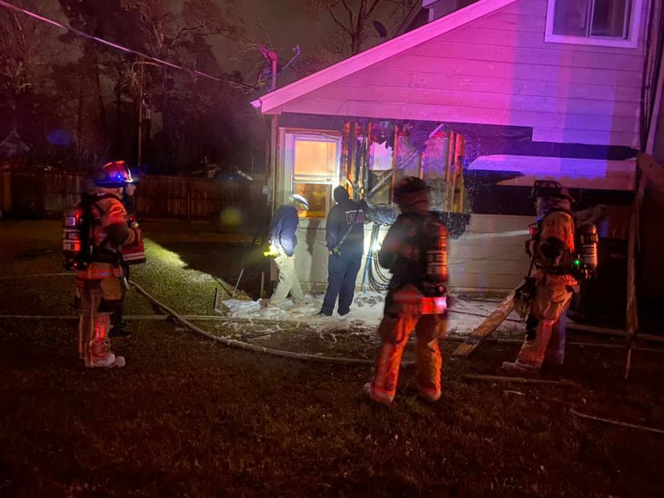 Firefighters in gear stand around the side of a house at night. Some hold hoses, and others appear to survey damage near a window. The scene is illuminated by red and purple lights. The ground is wet, suggesting firefighting efforts.