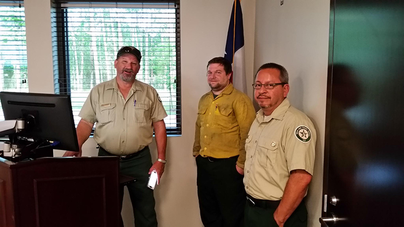 Three men in uniform stand in an office. Two wear beige shirts with patches, and one has a yellow shirt. They are next to a podium and computer. A window with blinds and a flag are in the background. They appear to be in a meeting or presentation.
