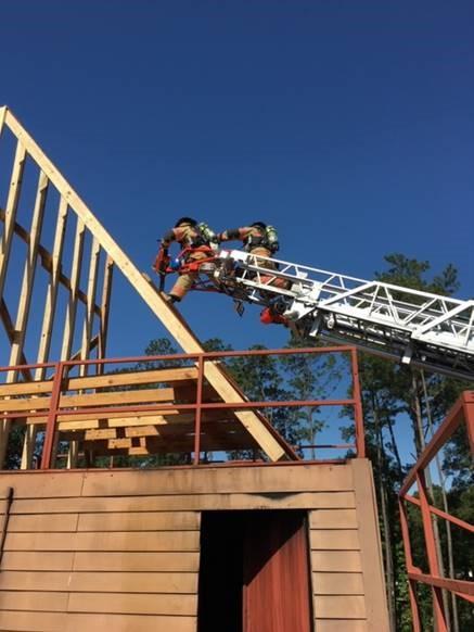 Two firefighters using a ladder truck spray water onto the frame of a wooden building with a clear blue sky in the background.
