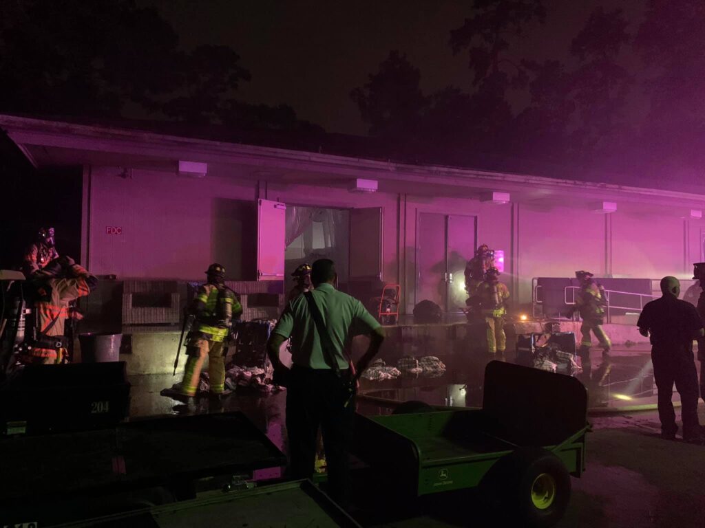 Firefighters work at night outside a building with thick smoke, illuminated by pink and purple lights. Several personnel in full gear are present, with carts and equipment visible in the foreground.