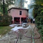 A fire-damaged house emits smoke, with firefighters present. Hoses spread across the driveway, and foam covers parts of the ground. Trees and bushes border the scene, under a partly cloudy sky.