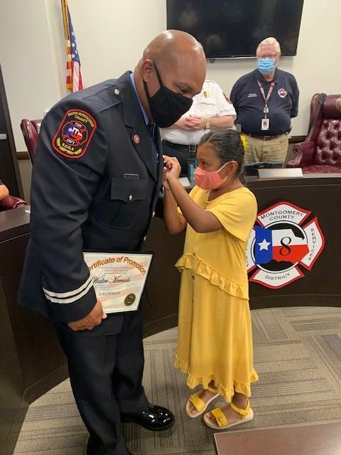 A firefighter in uniform receives a badge from a young girl in a yellow dress. Both are wearing masks. The firefighter holds a certificate. Other people in uniform are in the background in a meeting room.