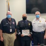 Three men in uniform stand indoors. The man in the center holds a certificate of promotion, and all are wearing face masks. An American flag is visible in the background.