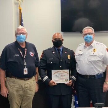 Three men in uniform stand indoors. The man in the center holds a certificate of promotion, and all are wearing face masks. An American flag is visible in the background.