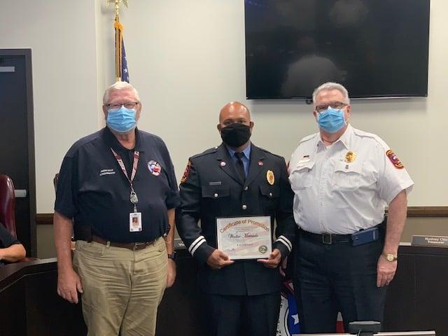 Three men in uniform stand indoors. The man in the center holds a certificate of promotion, and all are wearing face masks. An American flag is visible in the background.