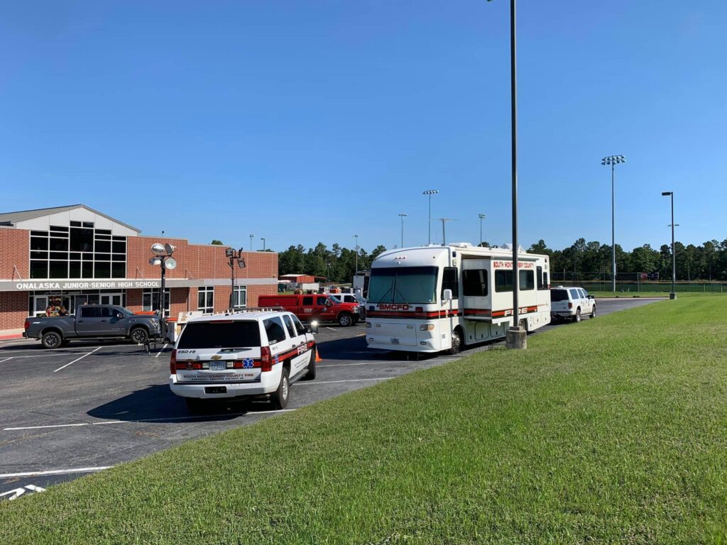 A parking lot with three emergency vehicles and a large mobile unit near a brick building. The sky is clear and blue. There are trees and a sports field with light poles in the background.