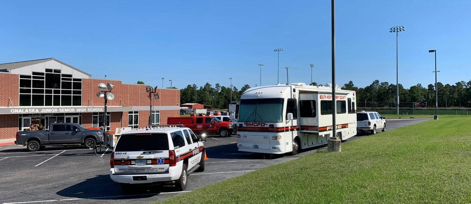 A parking lot with three emergency vehicles and a large mobile unit near a brick building. The sky is clear and blue. There are trees and a sports field with light poles in the background.