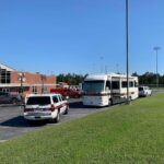 A parking lot with three emergency vehicles and a large mobile unit near a brick building. The sky is clear and blue. There are trees and a sports field with light poles in the background.