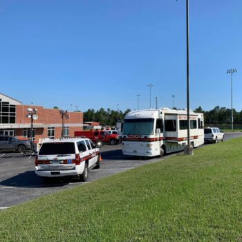 A parking lot with three emergency vehicles and a large mobile unit near a brick building. The sky is clear and blue. There are trees and a sports field with light poles in the background.