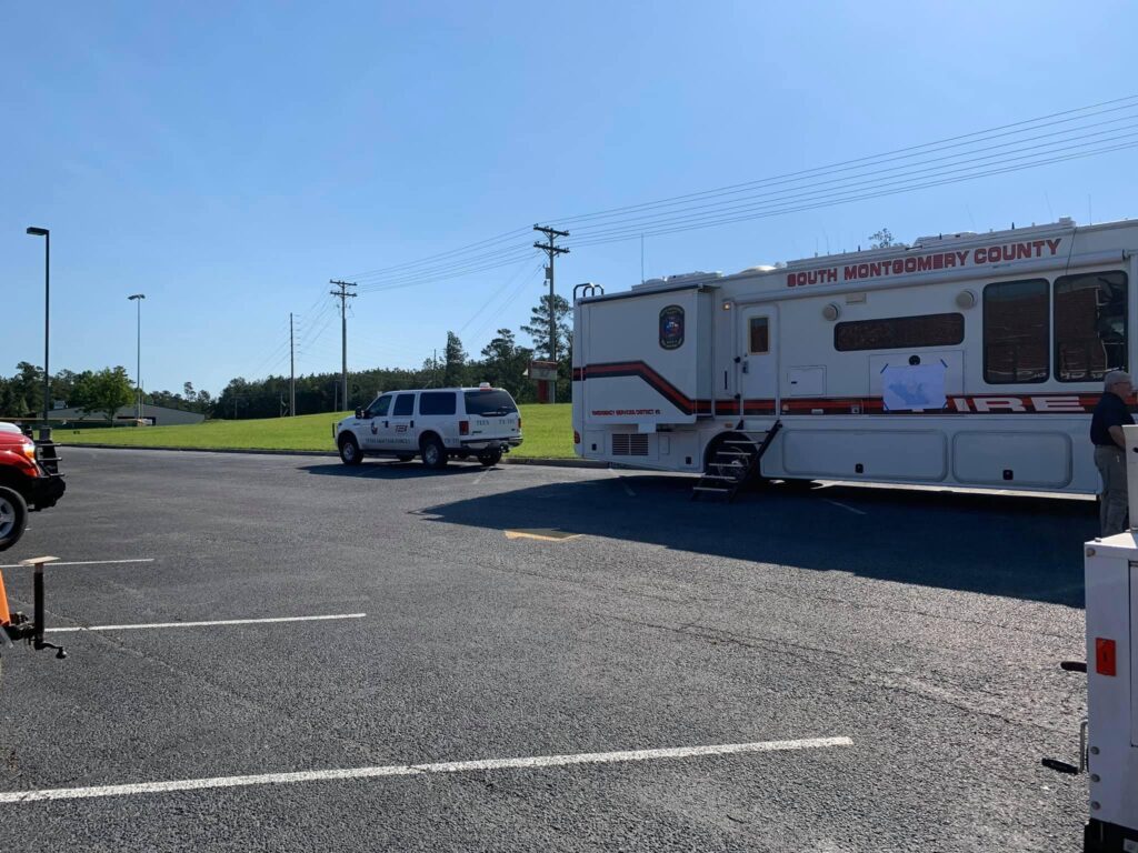 An emergency response vehicle labeled South Montgomery County Fire is parked on an asphalt surface. A white SUV with a similar logo is nearby. Its a sunny day, and the area is surrounded by grassy fields and power lines.