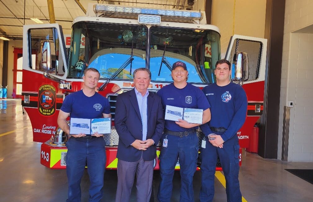 Four men stand in front of a fire truck inside a station. Three wear firefighter uniforms holding what appear to be certificates, and one is in a suit. The fire truck is red and white with visible logos and the number 113.