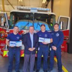 Four men stand in front of a fire truck inside a station. Three wear firefighter uniforms holding what appear to be certificates, and one is in a suit. The fire truck is red and white with visible logos and the number 113.