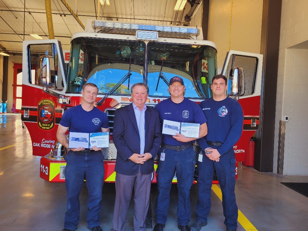 Four men stand in front of a fire truck inside a station. Three wear firefighter uniforms holding what appear to be certificates, and one is in a suit. The fire truck is red and white with visible logos and the number 113.