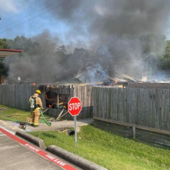 A firefighter in protective gear stands near a wooden fence, spraying water toward a house engulfed in thick black smoke. A red fire truck is partially visible on the left, and a stop sign is in the foreground.