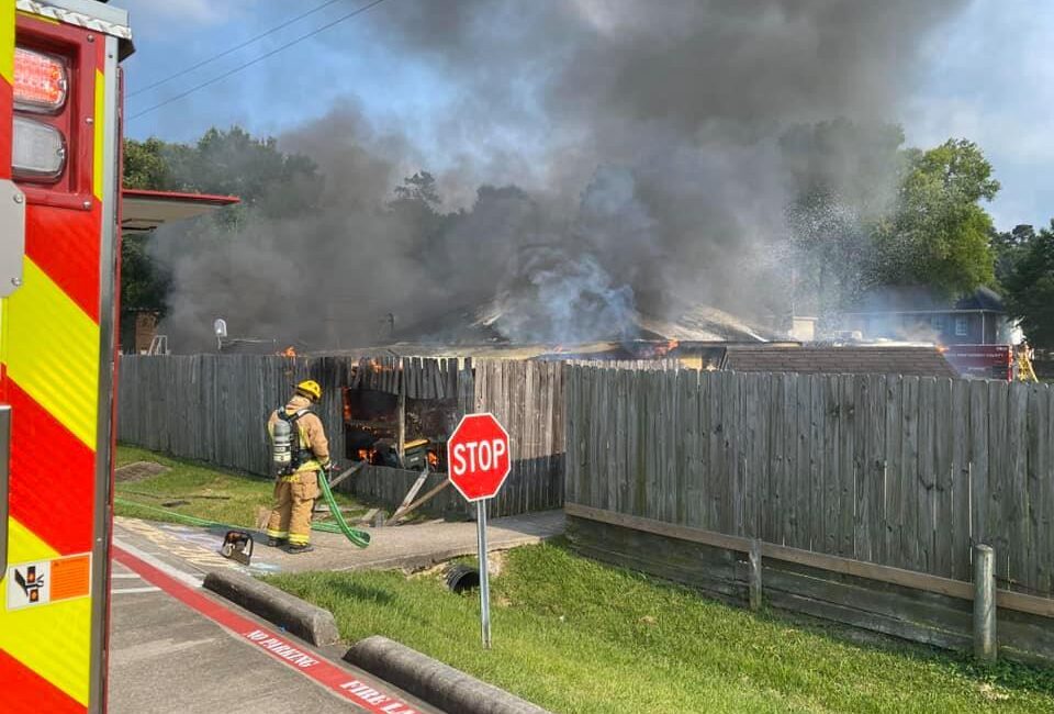 A firefighter in protective gear stands near a wooden fence, spraying water toward a house engulfed in thick black smoke. A red fire truck is partially visible on the left, and a stop sign is in the foreground.