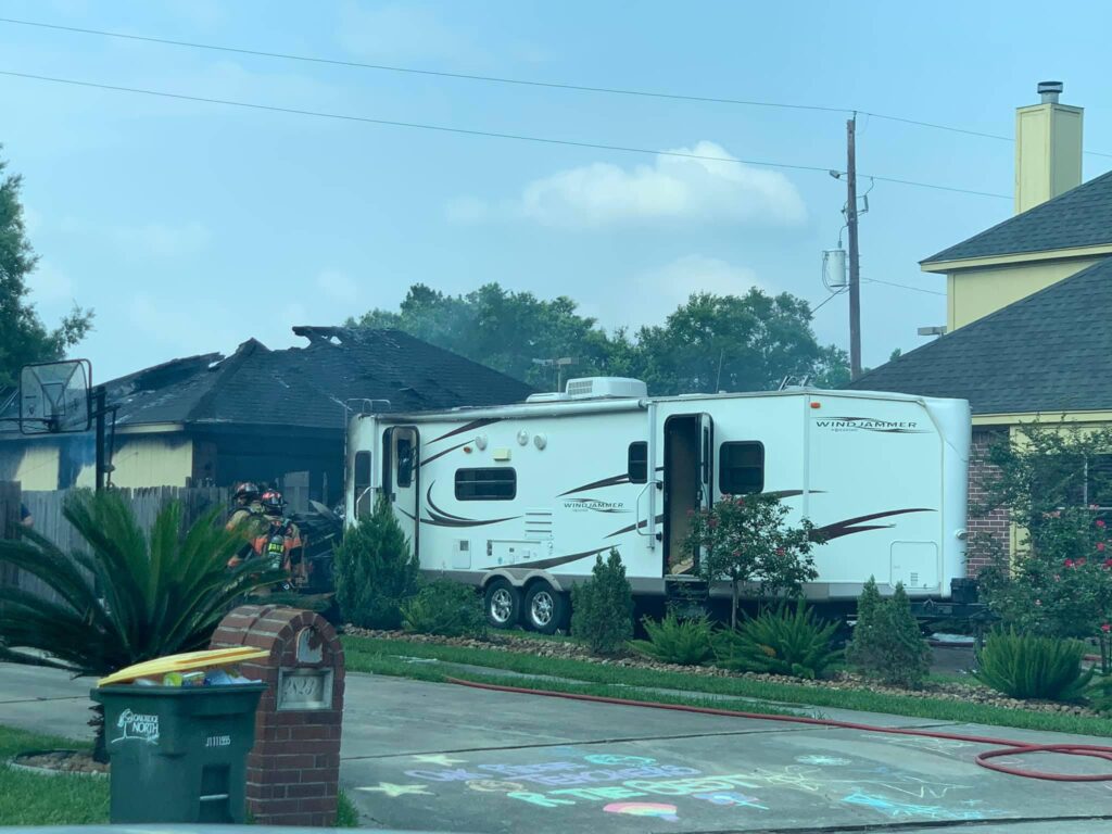 Firefighters extinguish a house fire next to a white RV parked in a driveway. A hose is laid out, and theres a basketball hoop nearby. The garage and part of the house are heavily damaged. A sidewalk shows chalk drawings.