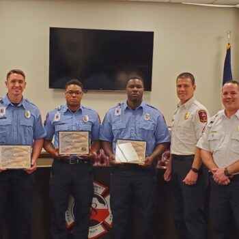 A group of seven uniformed individuals stands together in a room with flags and plaques behind them. Three in blue uniforms are holding certificates. Theyre standing in front of a large monitor and fire department insignia.