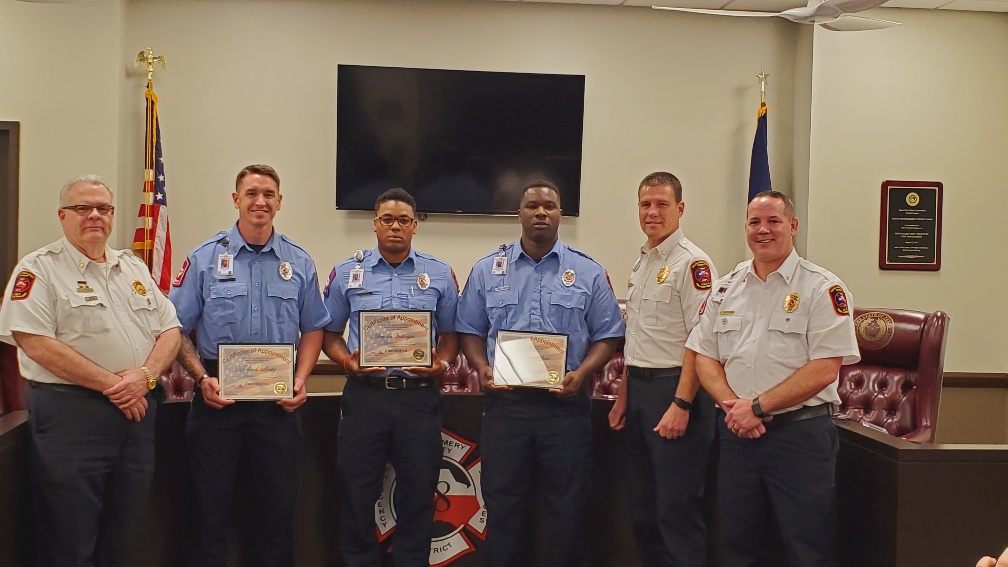 A group of seven uniformed individuals stands together in a room with flags and plaques behind them. Three in blue uniforms are holding certificates. Theyre standing in front of a large monitor and fire department insignia.