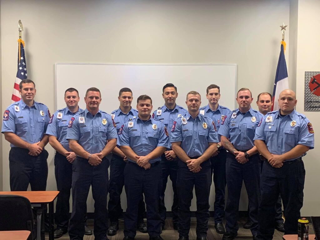 Eleven people in matching blue uniforms pose in two rows inside a room. American and another flag are visible behind them. They stand in front of a whiteboard and desks are in the foreground.