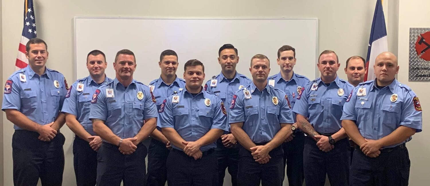 Eleven people in matching blue uniforms pose in two rows inside a room. American and another flag are visible behind them. They stand in front of a whiteboard and desks are in the foreground.