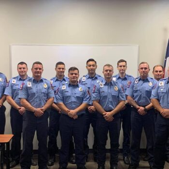Eleven people in matching blue uniforms pose in two rows inside a room. American and another flag are visible behind them. They stand in front of a whiteboard and desks are in the foreground.