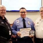 Three men in a room, two wearing white shirts with badges and one in a blue uniform holding a Certificate of Promotion. They stand in front of a screen displaying text on the wall, smiling at the camera.