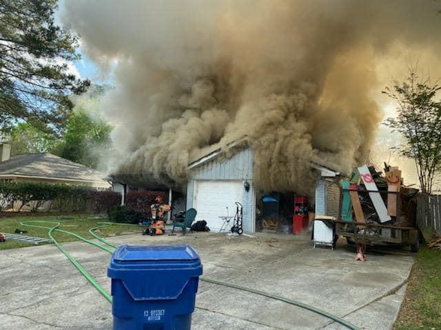 A suburban house with a garage is engulfed in thick, billowing smoke. Several hoses are laid out on the driveway, and a blue recycling bin is in the foreground. Various items are scattered near the garage entrance.