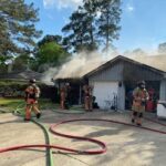 Firefighters in protective gear work to extinguish a house fire. Smoke rises from the roof and garage. Several hoses are stretched across the driveway. Trees and a clear sky are visible in the background.