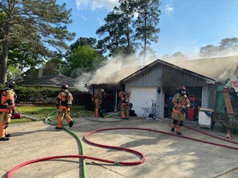 Firefighters in protective gear work to extinguish a house fire. Smoke rises from the roof and garage. Several hoses are stretched across the driveway. Trees and a clear sky are visible in the background.